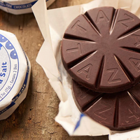 Two round dark chocolate discs, the traditional Mexican format, with segmented starburst patterns and embossed Taza Chocolate branding, resting on white paper over a rustic wooden surface. Part of a wrapped round labeled 'Salt' is visible on the left indicating the sea salt range